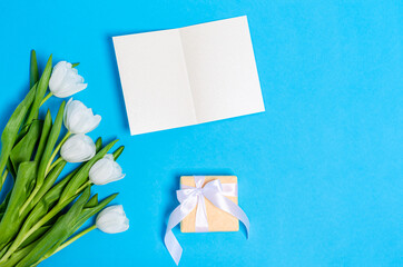 Bouquet of white tulips with a greeting card and a gift box on a light blue background. Flat lay, copy space, spring concept Top view.