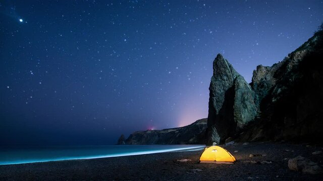 Glowing camping tent on a sea shore with rocks at night under the twinkling starry sky