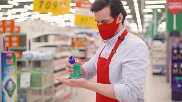 Handsome Caucasian Young Seller Man Wears Red Face Mask And Apron Cleaning Hands With Sanitizer Gel In Supermarket. Male Employee Using Alcohol Sanitiser Before Working To Prevent Coronavirus Concept.