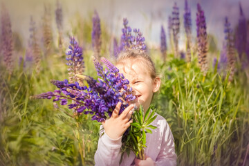 Beautiful little girl holds a large bouquet of purple lupins in flowering field. Flowers cover the girl's face. Blooming lupine flowers. Nature concept. Provence. Childhood. Summer vacation.