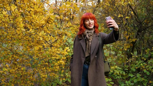 a girl with red hair in a coat, takes a selfie against the background of autumn foliage in the park and then posts it to the network