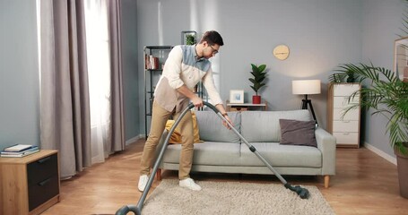 Caucasian handsome serious young man vacuuming carpet cleaning modern apartment, clean up, housekeeper, guy doing domestic tasks and cleans living room using vacuum cleaner, householding