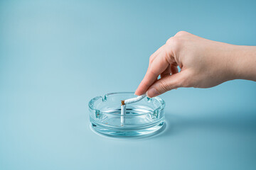 A hand breaks a cigarette in an ashtray on a blue background. Side view with copy space. The concept of the dangers of smoking.