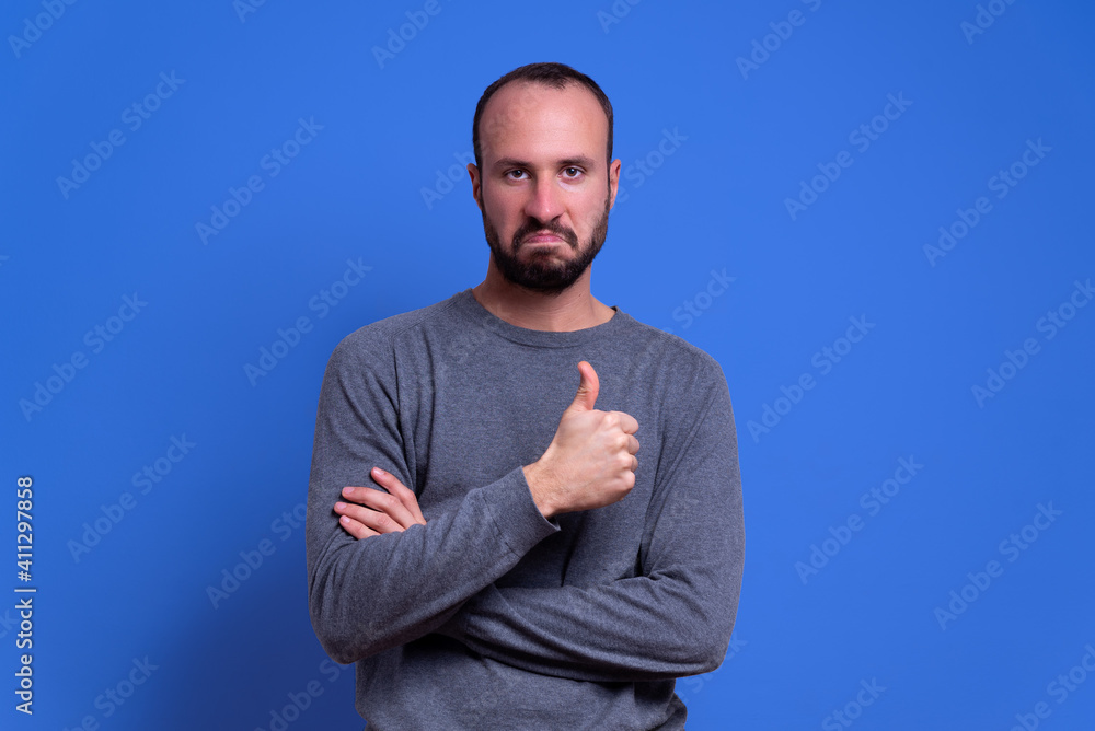 Wall mural young man with beard and casual clothing giving thumbs up as a gesture of approval with serious and convinced expression on blue background. expression of pride and winner