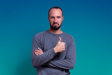 young man with beard and casual clothing giving thumbs up as a gesture of approval with serious and impressed expression on blue fading background. expression of pride and winner - obrazy, fototapety, plakaty