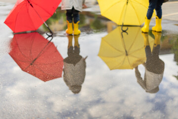 Yellow children's rubber boots on the street next umbrellas as a background. Reflection in the water in a puddle. Legs of two children stand together after rain