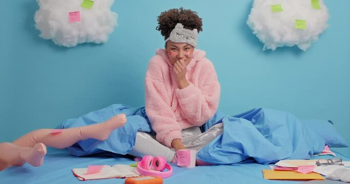 Mysterious surprised woman indicates at inflated doll feels lonely while staying at home on self isolation wears soft pajama and sleepmask stays in bed with papers around undergoes beauty procedures