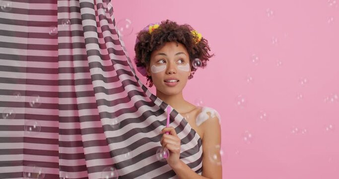 Dreamy satisfied Afro American woman enjoys taking shower holds tooth brush looks around with thoughtful cheerful expression poses behind curtain foam bubbles falling around. Personal care concept