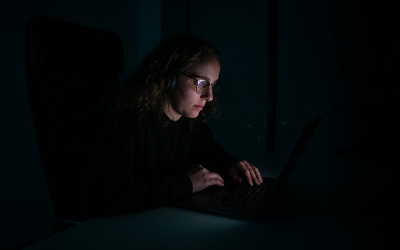 Woman with headphones typing on her laptop in the dark of the night. Overwork.