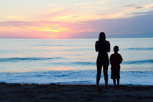 Mother and son watching sunrise - pink sky and calm, blue waters with silhouette of boy and girl 