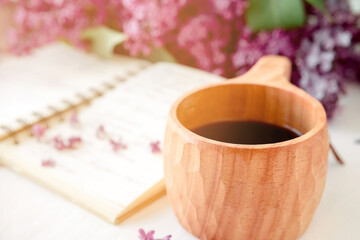 Wooden finnish cup with coffee on the table. Nearby lies a closed notebook for notes and a bouquet of lilacs. Good morning and good mood, planning a day at breakfast, concept..