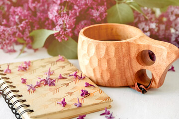 Wooden finnish cup with coffee on the table. Nearby lies a closed notebook for notes and a bouquet of lilacs. Good morning and good mood, planning a day at breakfast, concept.