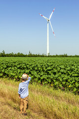 A child from a dirt road looks at wind turbines against a background of cloudy blue sky and green field
