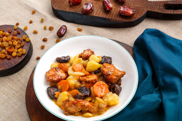 A dish of Jewish cuisine sweet tsimes with carrots, dates and turkey meat in a white plate on a round board on a linen background near candied fruits.