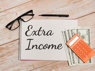 Phrase Extra Income written on  notebook with pen,calculator,fake money and eye glasses on wooden...