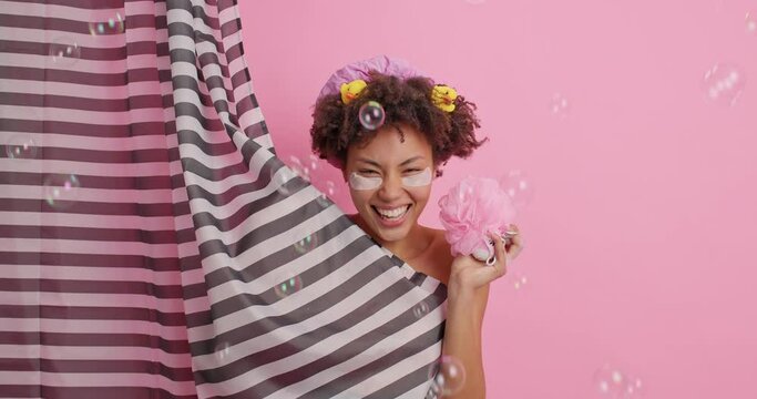 Funny dark skinned woman takes shower makes funny grimace pouts cheeks applies collagen patches under eyes hides behind curtain poses around flying bubbles. Body care and daily pampering concept