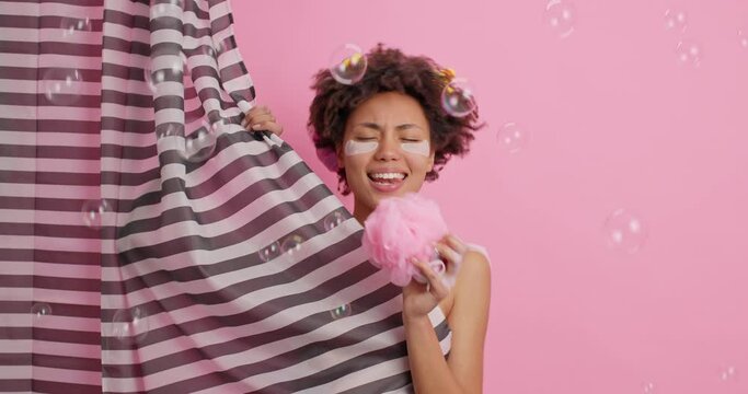 Carefree cheerful dark skinned woman with curly hair sings in shower sponge has fun while having douche poses behind striped curtain takes care of body. Skin care hygiene and refreshment concept