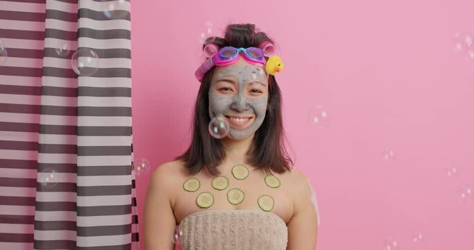 Pretty smiling eastern woman has gentle look at camera applies nourishing clay mask for reducing wrinkles feels refreshed after taking shower isolated over pink background. Asian female in douche