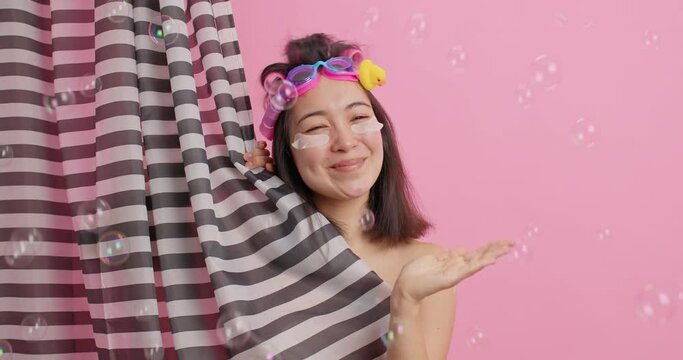 Spa and body washing concept. Happy young Asian woman keeps palm raised catches bubbles has good mood while taking shower isolated over pink background enjoys hygiene procedures at bathroom.