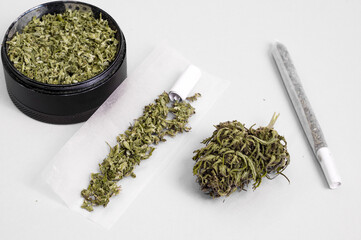 grinder full of dry herb and joint with medical marijuana with paper filter and piece of rolling paper on white background