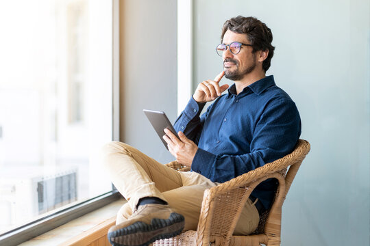 Casual business man wearing glasses sitting holding his tablet. Successful male portrait with thinking face expression working from home by the window