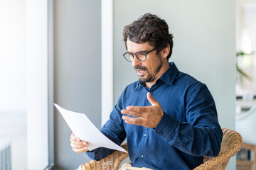 Casual man looking surprised reading bills to pay. Taxes, bank statement and loan debt. Male...