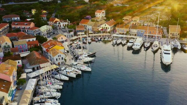 Drone flying backwards over the beautiful marina and town of Fiskardo in Greece. There are a lot of boats in the marina. It is a beautiful evening around sunset, with sun flares in right side of frame