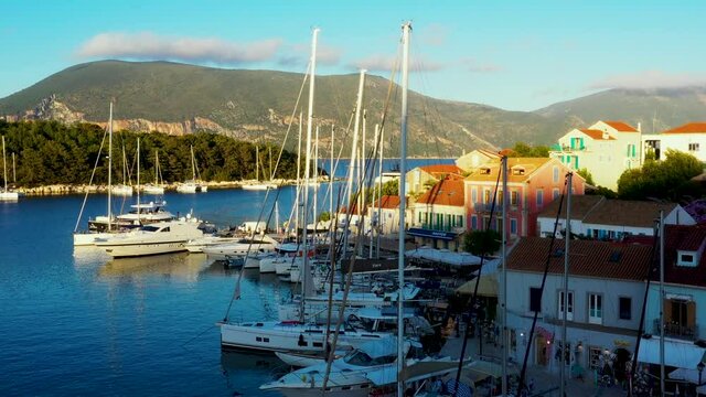 Drone taking off over the beautiful marina and town of Fiskardo in Greece. There are a lot of boats in the marina. It is a beautiful evening around sunset. There are mountains in the background. 4K