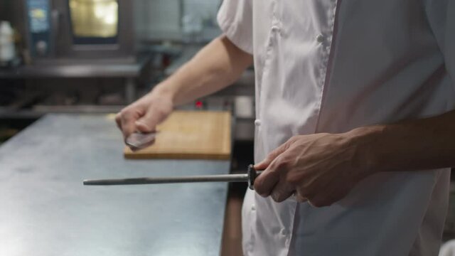 Midsection slowmo of unrecognizable chef in white jacket sharpening his knife t restaurant kitchen