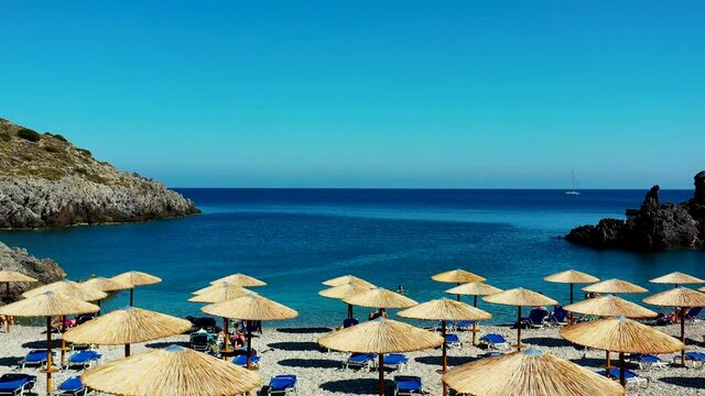 Drone footage of the Halkos Kythera beach in Greece. The drone is facing the clear blue ocean, and takes off over the parasols and sun beds.