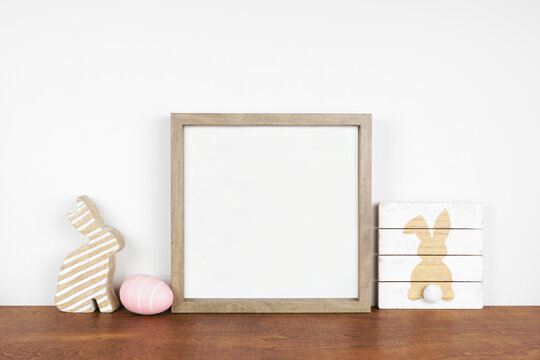 Mock up wood frame with Easter decor on a wood shelf. Shabby chic wood sign, egg, bunny. Square frame against a white wall. Copy space.