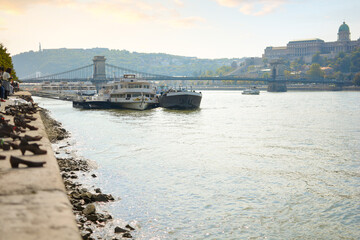 Fototapeta na wymiar Boats line the banks of the Danube River near the Shoes on the Danube memorial with the Chain Bridge and Royal Palace behind, in Budapest, Hungary
