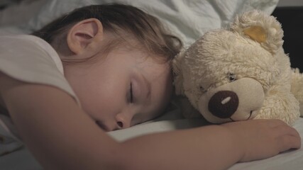 The kid sleeps at home on the sofa in the children's room. The sleeping baby is happy and carefree in bed, hugging a teddy bear toy. The mother covered her child with a blanket. Happiness in dream.