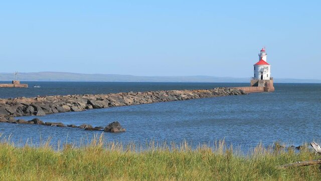 Lighthouse with red roof at end of a rock pier and a fishing boat on the blue water, on a sunny day, with nearby grass swaying gently in the breeze.