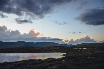 Scenic mountain scenery with lilac reflection of sunset in lake. Colorful mountain landscape with illuminating color in violet clouds in dawn gradient sky. Beautiful view to sunrise and lilac lake.