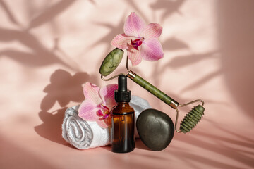 Obraz na płótnie Canvas Photo for the spa salon. Roller and Guasha scraper for facial massage, massage oil, black hot spa stones, pink orchids, white towel on a pink background. Balance and levitation trending style
