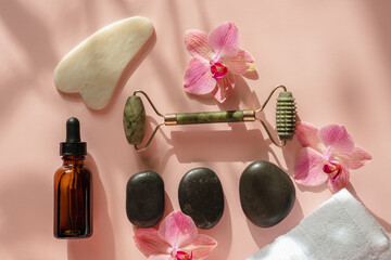 Obraz na płótnie Canvas Photo for the spa salon. Roller and Guasha scraper for facial massage, massage oil, black hot spa stones, pink orchids, white towel on a pink background.