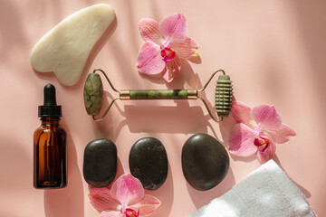 Photo for the spa salon. Roller and Guasha scraper for facial massage, massage oil, black hot spa stones, pink orchids, white towel on a pink background. Close-up top view