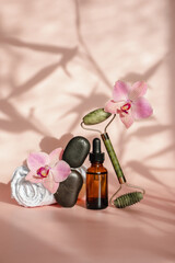 Photo for the spa salon. Roller and Guasha scraper for facial massage, massage oil, black hot spa stones, pink orchids, white towel on a pink background. 
Balance and levitation trending style