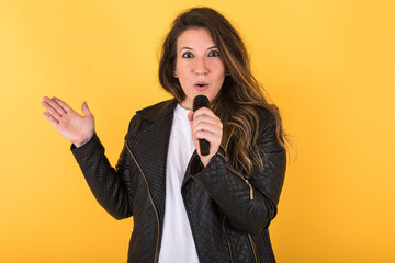 Young singer girl, wearing black leather jacket and microphone, making surprise gesture on yellow background.
