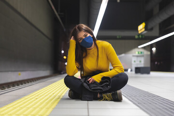 joven y atractiva muchacha con camisa amarilla y mascarilla azul para el coronavirus espera el metro en la parada con paciencia. - obrazy, fototapety, plakaty