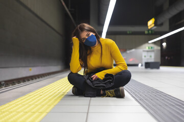 joven y atractiva muchacha con camisa amarilla y mascarilla azul para el coronavirus espera el metro en la parada con paciencia.