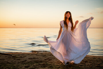 Fototapeta na wymiar Beautiful young woman in a white long dress near the sea at sunset stands