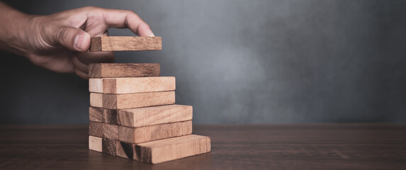 Close-up hand is placing wood block tower stacked in pyramid shape with caution to prevent collapse...