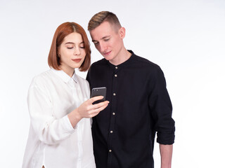 Girl shows guy screen of smartphone. Young couple with a phone in their hands. Portrait of a young couple on a white background. Students dressed in casual style stand side by side.
