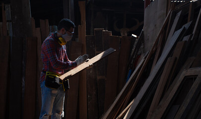 Yong Caucasian male carpenter in safety goggles, face mask, and ear muffs around his neck standing holding a wooden plank with looking, checking surface and quality of a wooden plank in a wood factory