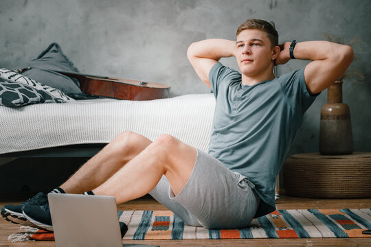 Young man goes in for sports at home, training online. The athlete makes the press, smiles, there is an open laptop, bed, vase, carpet nearby.