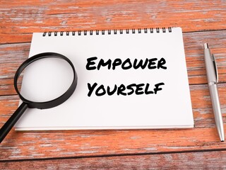 Top view of pen, magnifying glass and notebook written "EMPOWER YOURSELF" isolate on a wooden background.