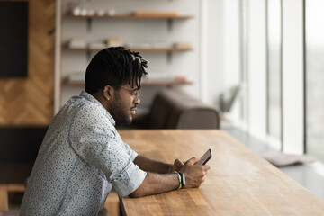 Side shot of happy afro american guy sit at desk read write text message on phone screen shop at...