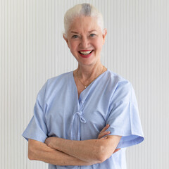 Senior older caucasian woman in hospital patient dress with positive smile face cross arms and...